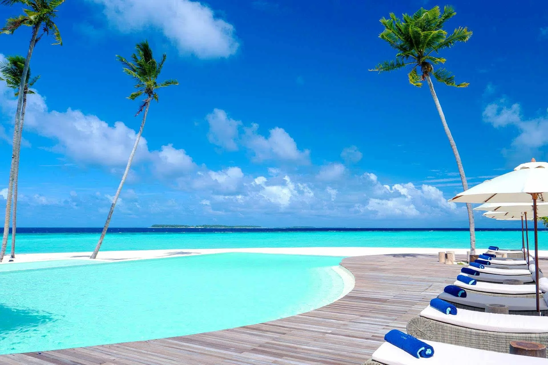 Poolside view with lounge chairs and umbrellas overlooking turquoise ocean and clear blue sky with scattered clouds, palm trees on the decking.