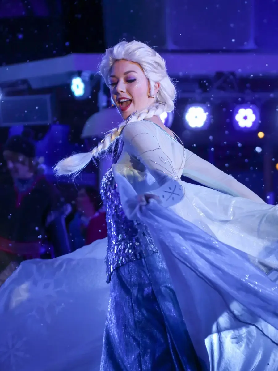 A woman dressed as Elsa from Frozen, performing at a winter-themed event with snow and glowing lights in the background.
