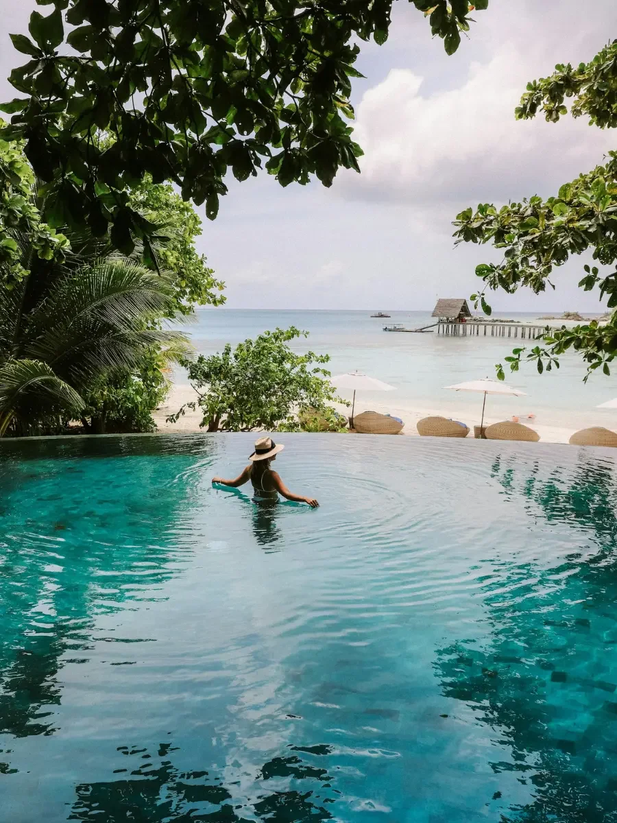 A woman relaxing in an infinity pool overlooking a beach with umbrellas, palm trees, and a pier with a hut, under overcast skies.