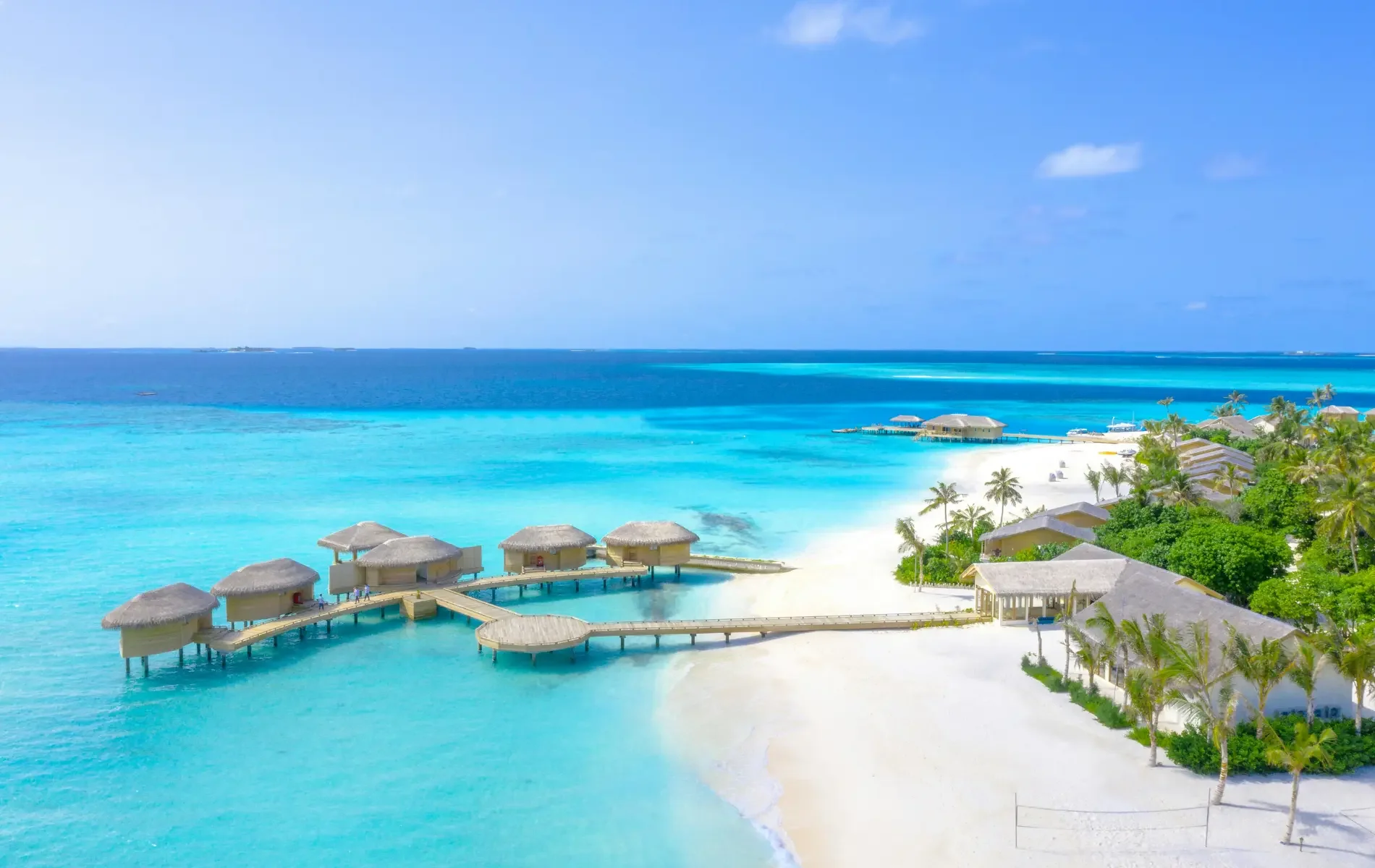 Aerial view of a tropical beach resort with overwater bungalows, white sandy beach, lush green palm trees, and clear turquoise water under a blue sky.