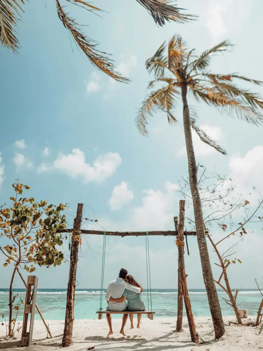 A couple sitting on a wooden swing on a sandy beach, with palm trees and the ocean in the background under a partly cloudy sky.