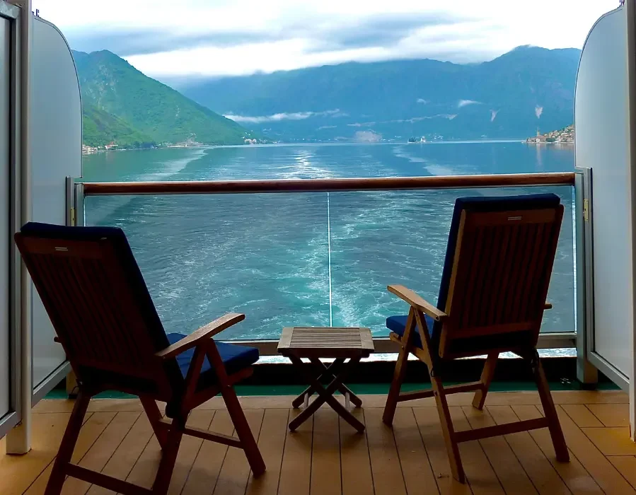 Two wooden chairs with blue cushions facing a lake view with mountains and cloudy sky, on a boat or ferry deck.