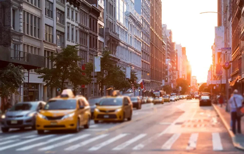 New York city street scene at sunset with yellow taxis, cars, and pedestrians. Tall buildings line both sides of the street.