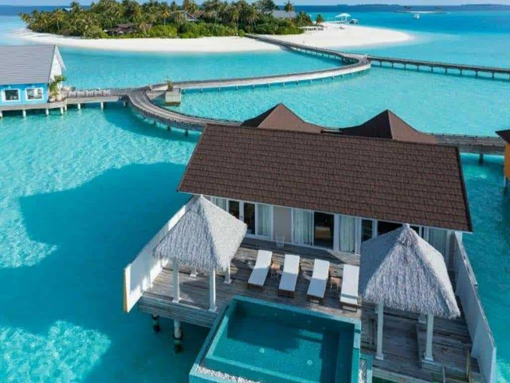 Overwater bungalow with thatched roofs, a small pool, and a deck with lounge chairs, located in clear blue water near a tropical island.