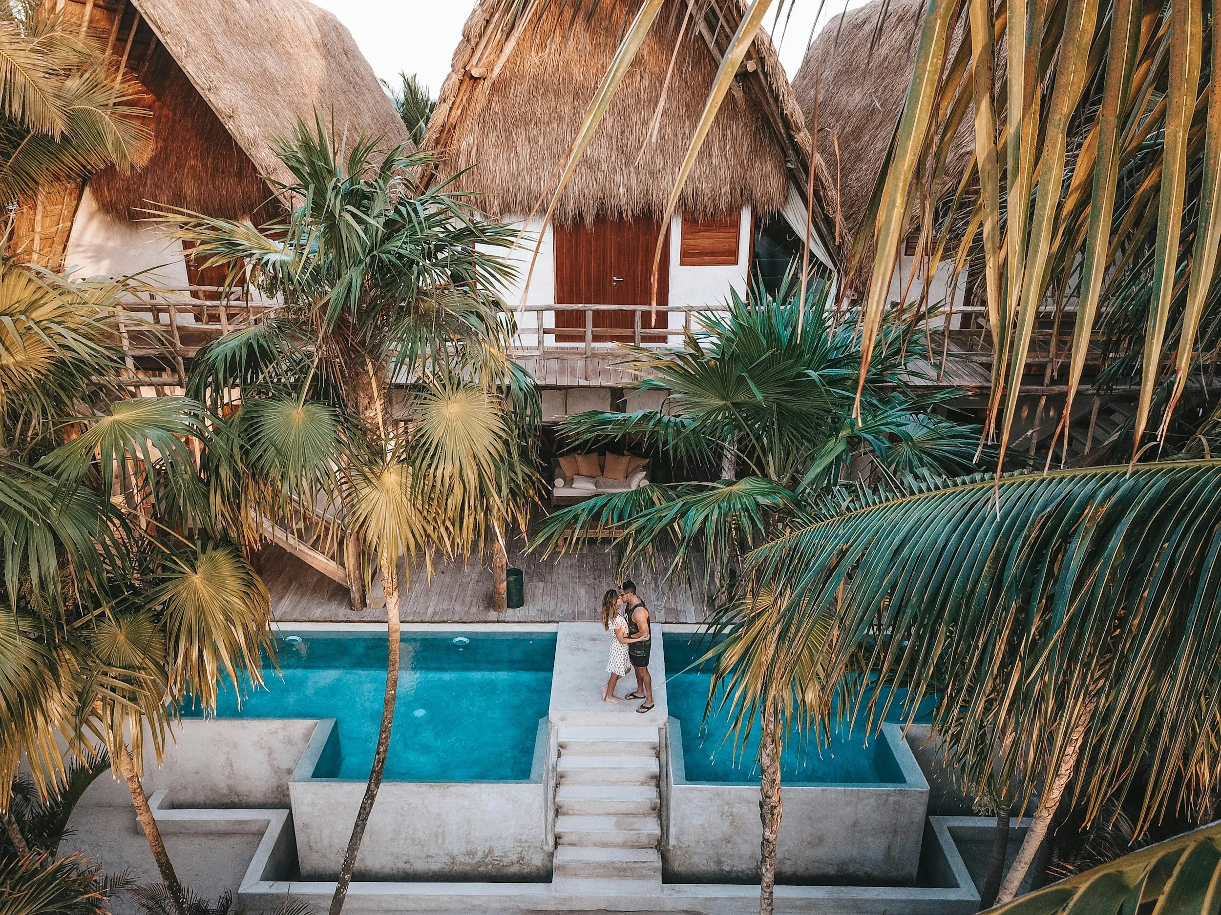 A couple standing on a small outdoor platform between two small pools, embracing, surrounded by lush tropical plants, with thatched-roof cottages in the background.