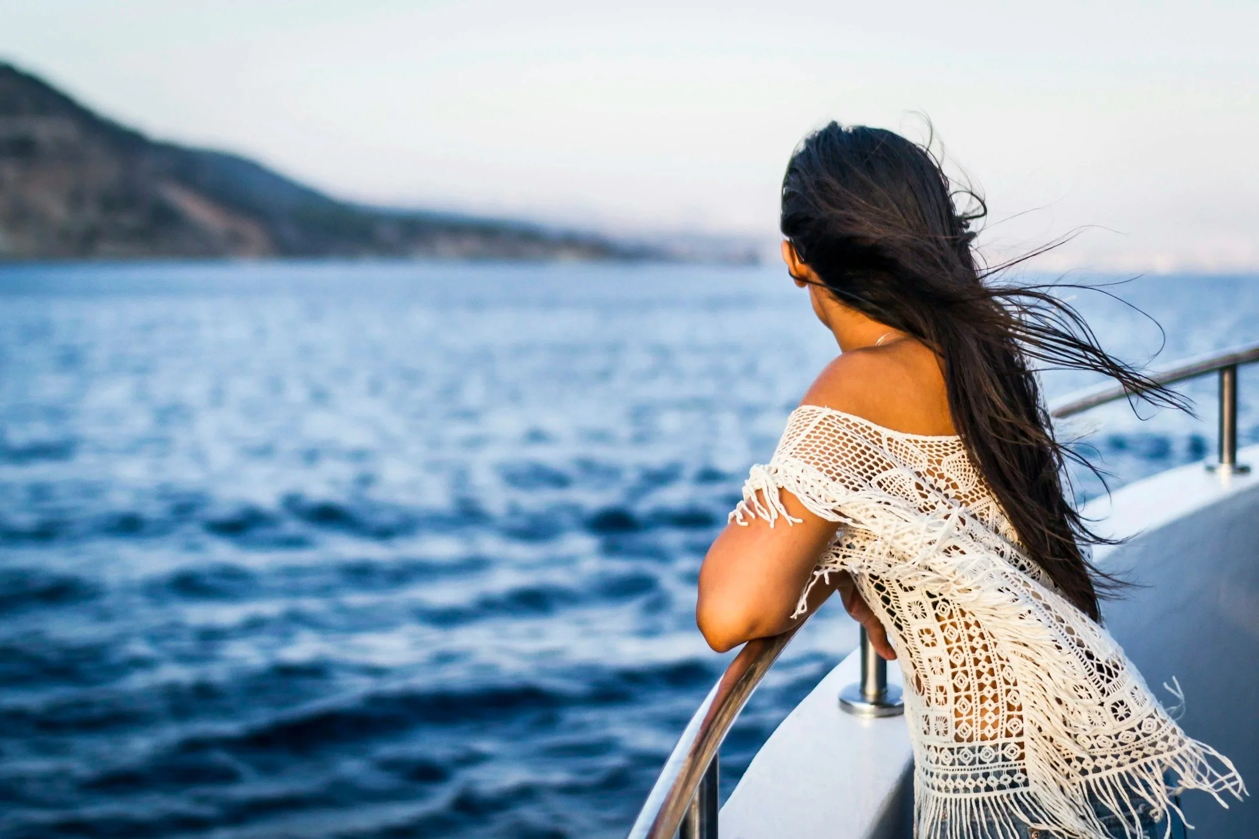 A woman with long black hair leaning on the railing of a cruise ship, looking out over the water with a distant landscape in the background.