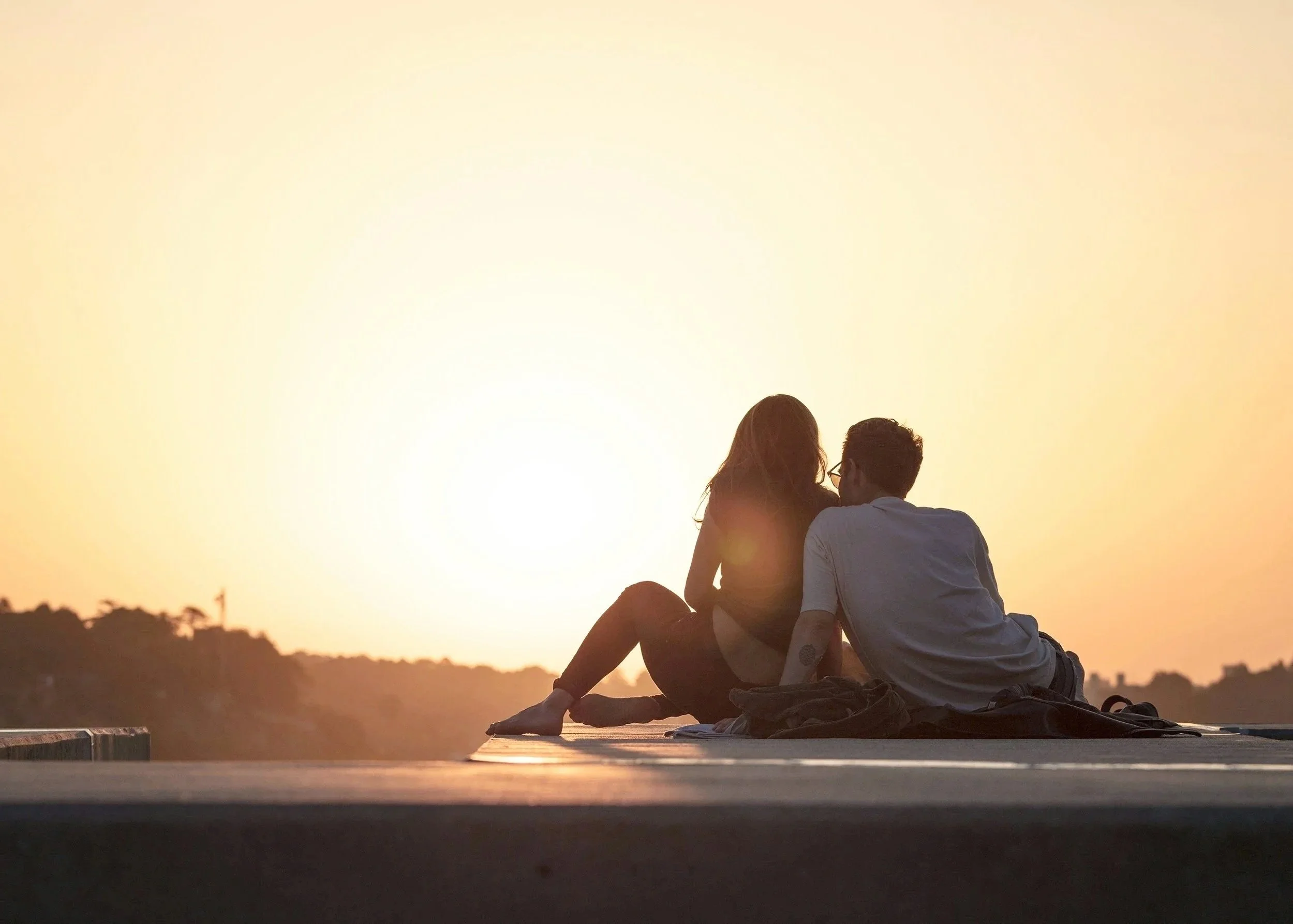 A couple sitting closely together on a raised platform, watching the sunset with warm light and a distant mountain range.