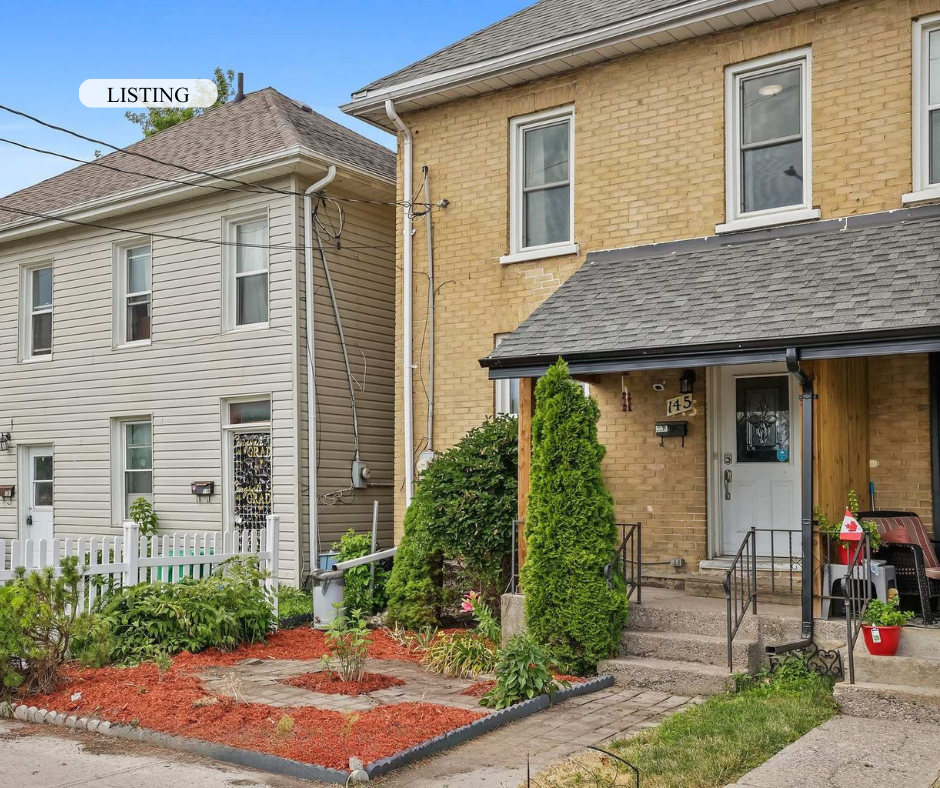 Front view of a yellow brick house with a porch, green bushes, and a small garden in front. To the left, part of a neighboring white house is visible. The house has steps leading to the front door, with a Canada flag and some patio furniture on the p