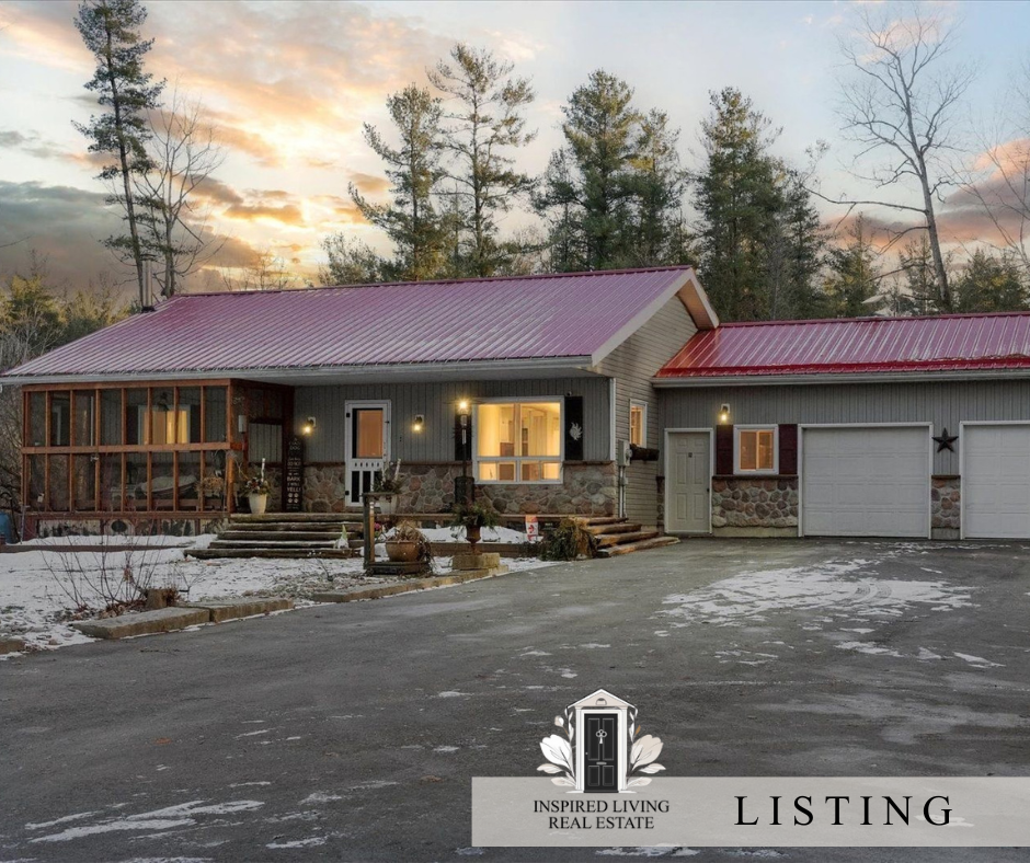 A house at sunset with a red metal roof, stone and siding exterior, a garage, front steps, and a porch with lighting, surrounded by tall trees and snow on the ground.