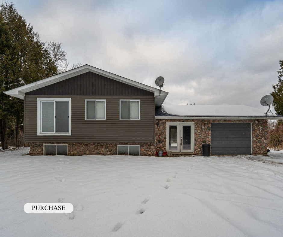 A house with a stone foundation and gray siding in a snowy landscape, with satellite dishes on the roof and a cloudy sky above.