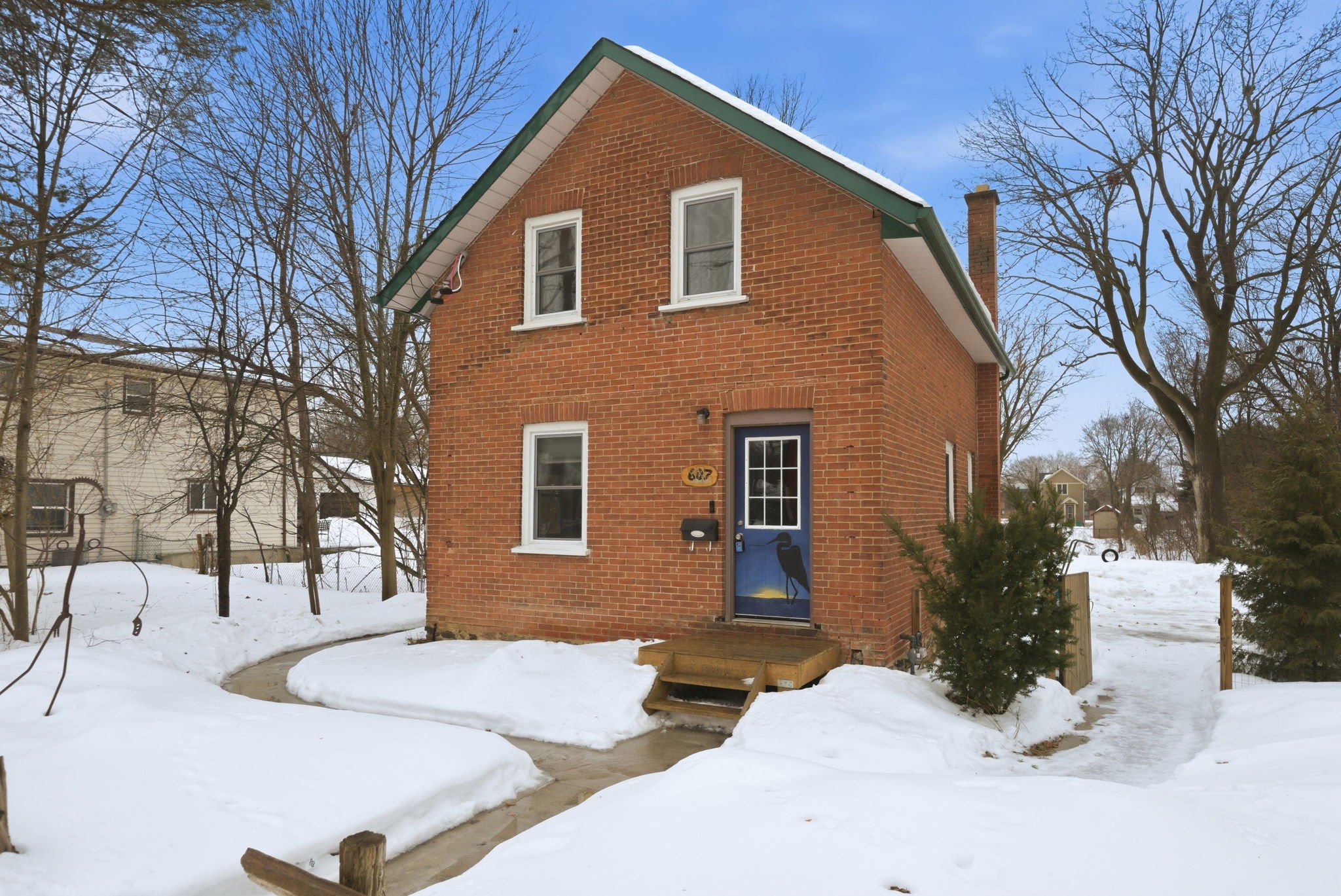 A brick house with a blue door featuring a heron silhouette, surrounded by snow, leafless trees, and a partly cloudy sky.