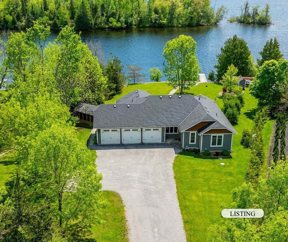 Aerial view of a house with a gray roof, surrounded by green trees, grass, and a body of water in the background.