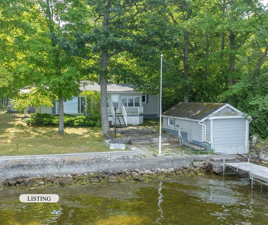 A lakeside property with a house, a garage, a flagpole, trees, and water with a dock.