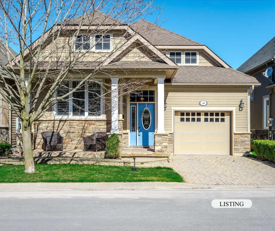A two-story house with beige exterior, stone accents, and a blue front door with an oval window, front porch with columns, a two-car garage, and a small tree in the front yard under a clear blue sky.
