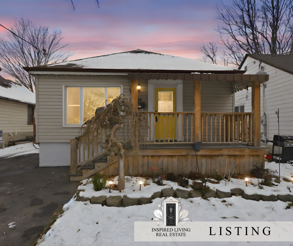 Front view of a small house with snow on the ground and a wooden front porch, illuminated by outdoor lights at dusk, with trees in the background.