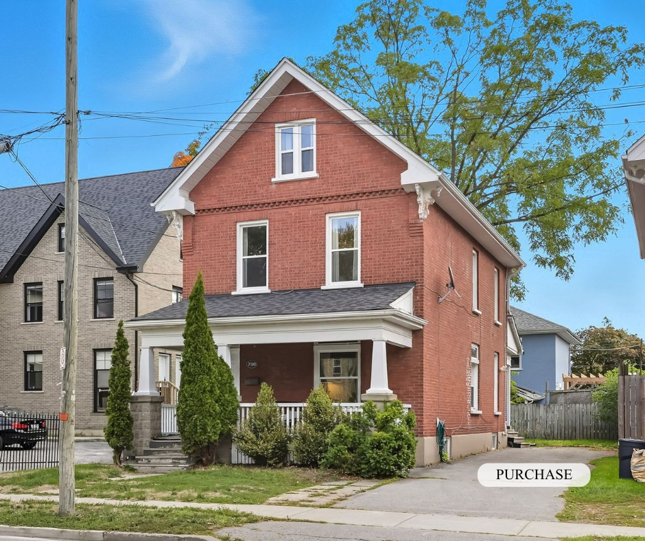 A large three-story red brick house with a white porch, surrounded by greenery and trees in a residential neighborhood.