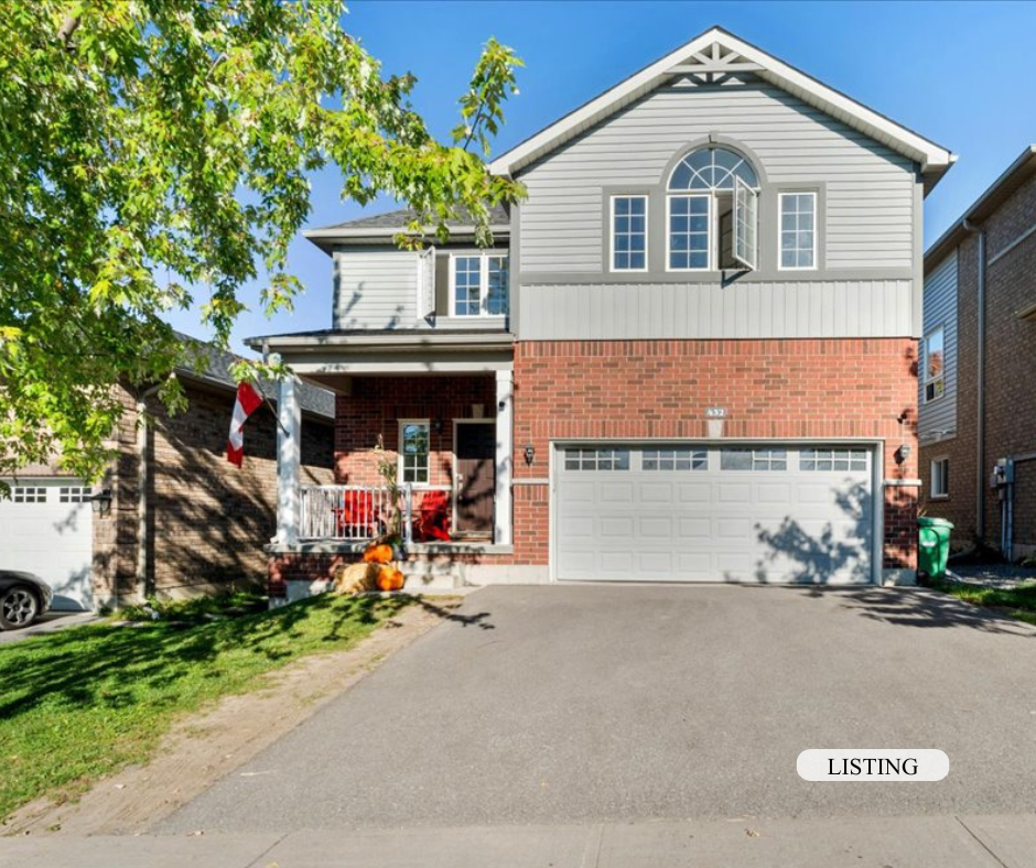 A two-story house with a grey upper level and a red brick lower level. It has a two-car garage, front porch with porch furniture, and a green lawn with tree shadows. There are pumpkins on the steps, suggesting fall decoration.