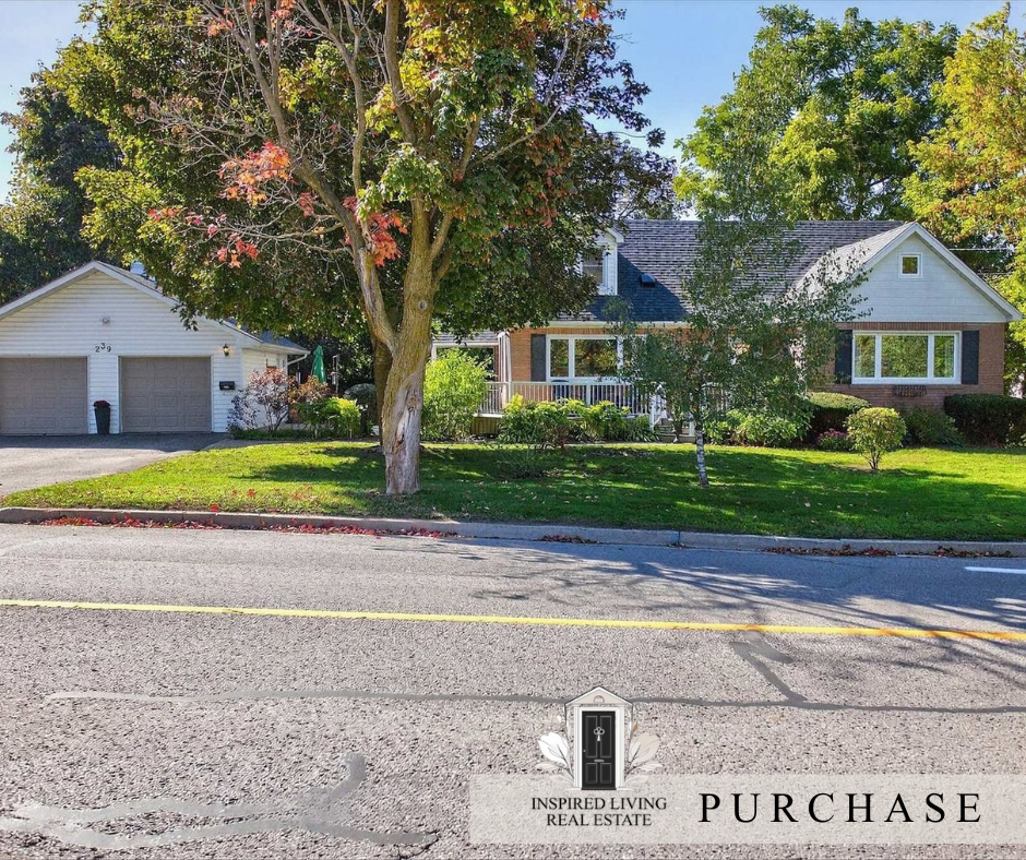A house with a front yard featuring a large tree, bushes, and a lawn. The house has a brick and white exterior with large windows and a porch. To the left is a detached garage with a driveway. The street in front is visible with a yellow dividing lin