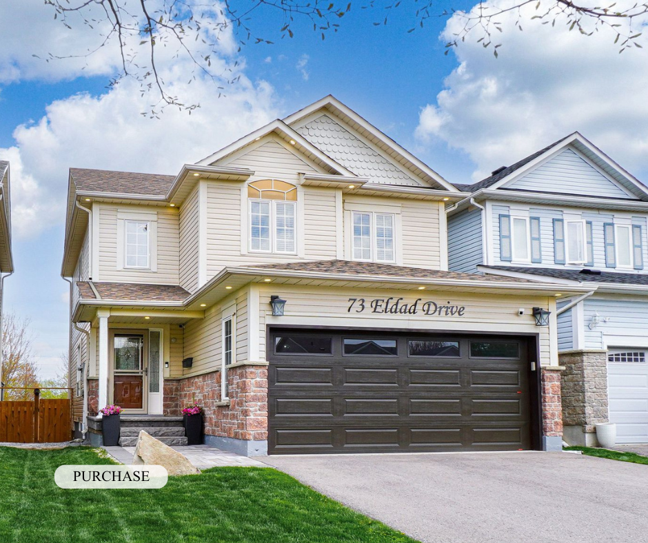Front view of a two-story house with beige siding, brick accents, and a large black garage door. The house has a small front porch with potted plants, and the address "73 Eldad Drive" is displayed above the garage. The lawn is well-maintained, and pa