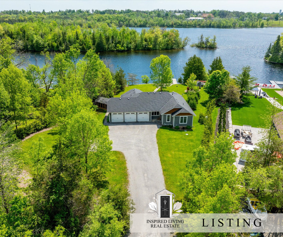 A house with a gray roof and white garage doors sits by a lake, surrounded by green trees and a well-maintained lawn.