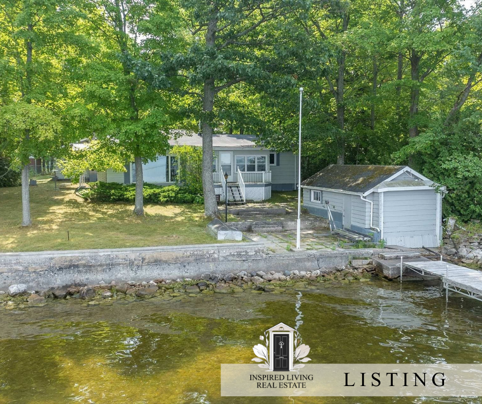 A lakeside property with a small dock, a gray house with a porch, and a detached garage surrounded by green trees.