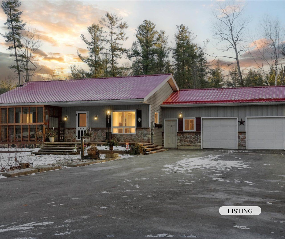 A house with a red metal roof, stone and siding exterior, surrounded by trees, with a driveway in front. There is snow on the ground and warm exterior lights are turned on inside and outside the house. The sky shows a sunset or sunrise.
