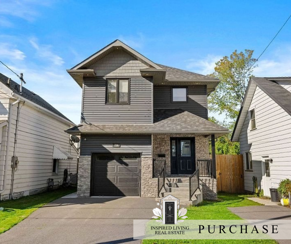 Two-story modern house with gray siding, stone accents, black garage door, front porch with stairs, and black front door. Residential neighborhood with houses on each side, green lawn, blue sky.