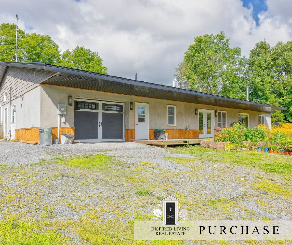 Single-story house with gray garage doors, surrounded by greenery, with a gravel driveway, garden beds, and a wooden deck, under a partly cloudy sky.
