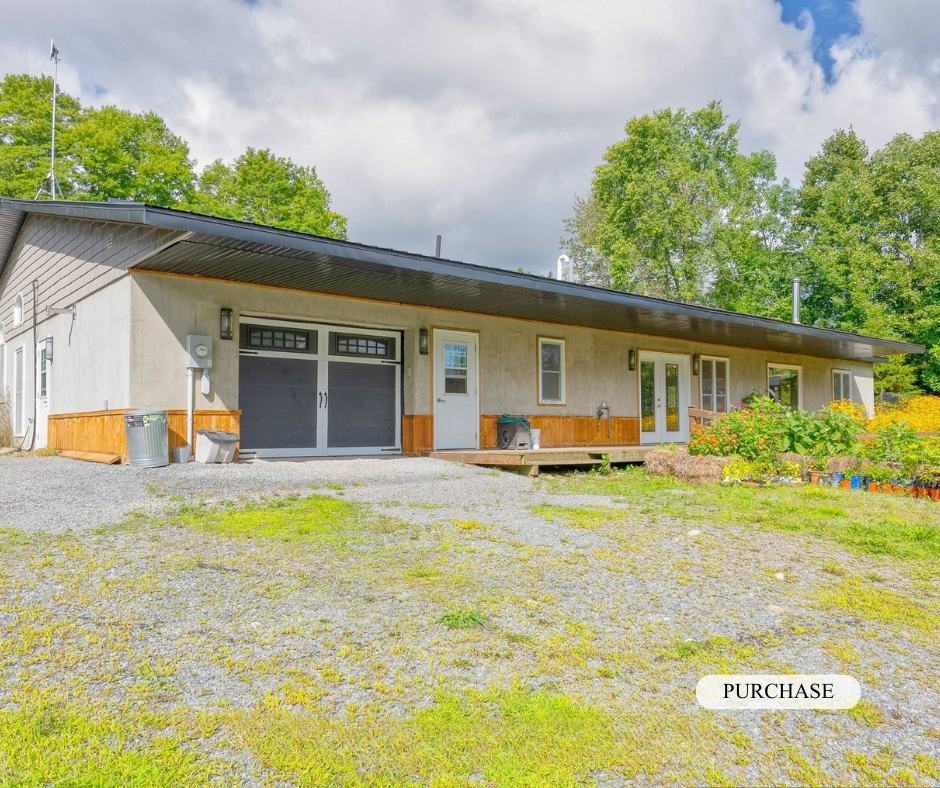 A single-story house with a sloped roof, white walls, and wooden siding at the bottom. There are white-framed windows, a white garage door, and a small deck in front. The yard has gravel and grass, with colorful flowers and plants to the right. Tall 