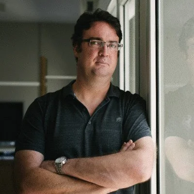 A man with glasses stands near a window, looking at the camera with arms crossed.