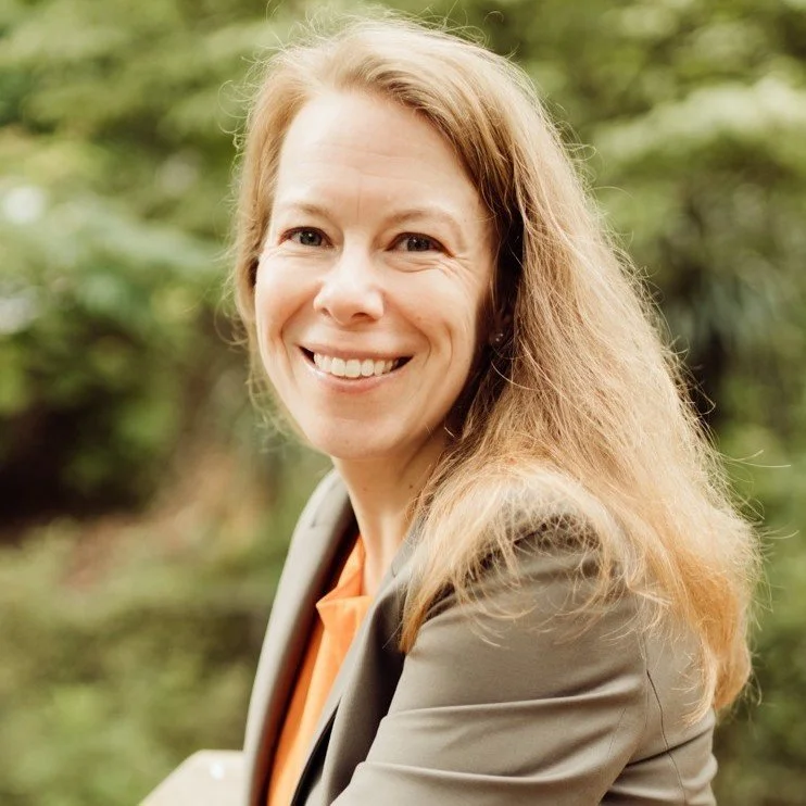 A woman with long blonde hair smiling outdoors with green foliage in the background.