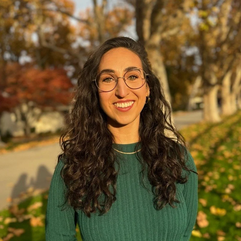 Young woman with long, curly dark hair wearing glasses and a green sweater, smiling outdoors in autumn with colorful trees in the background.