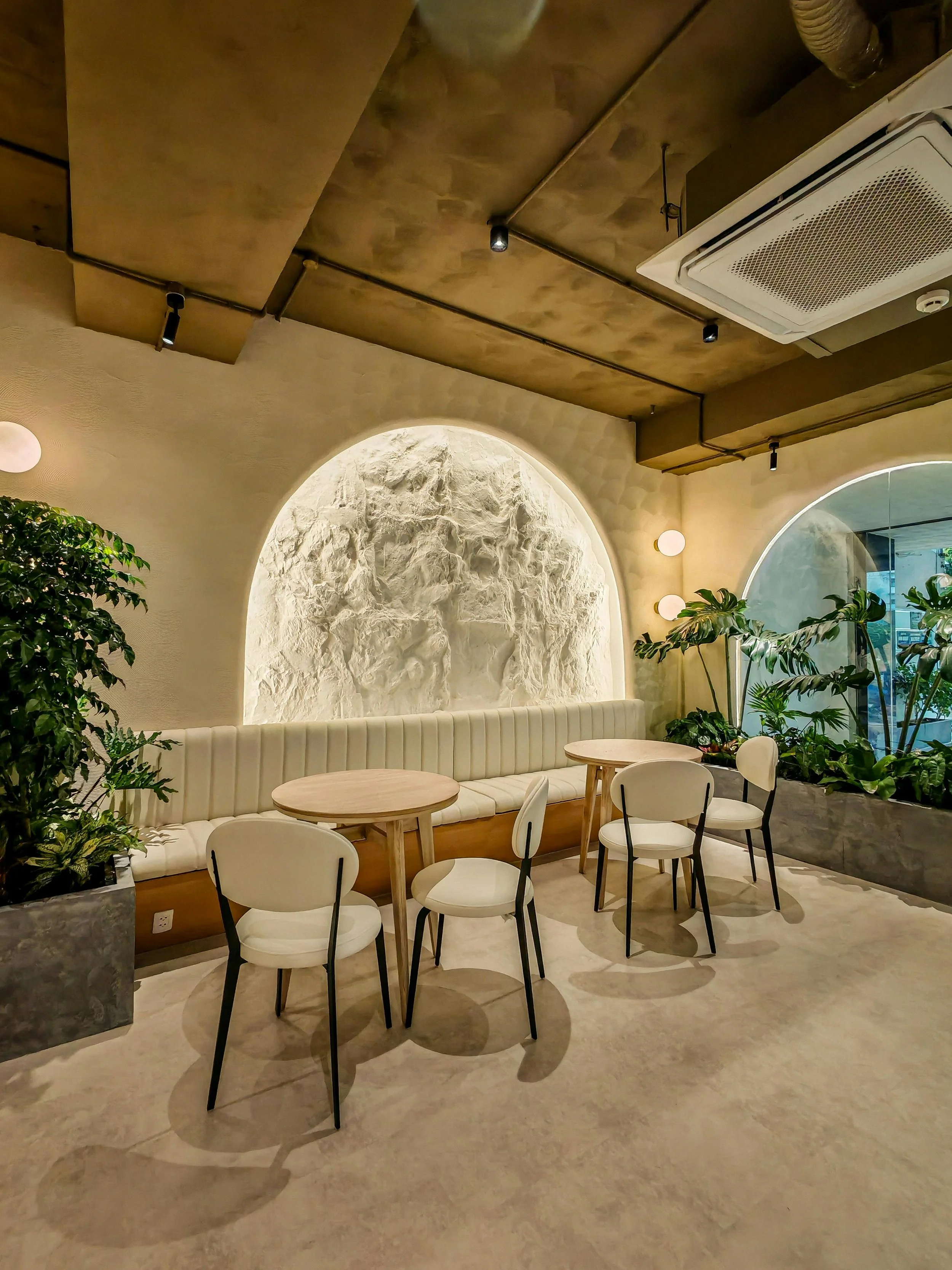 A modern indoor seating area with a built-in cushioned bench, surrounded by small round tables and white chairs, with green plants and decorative lighting on the walls.