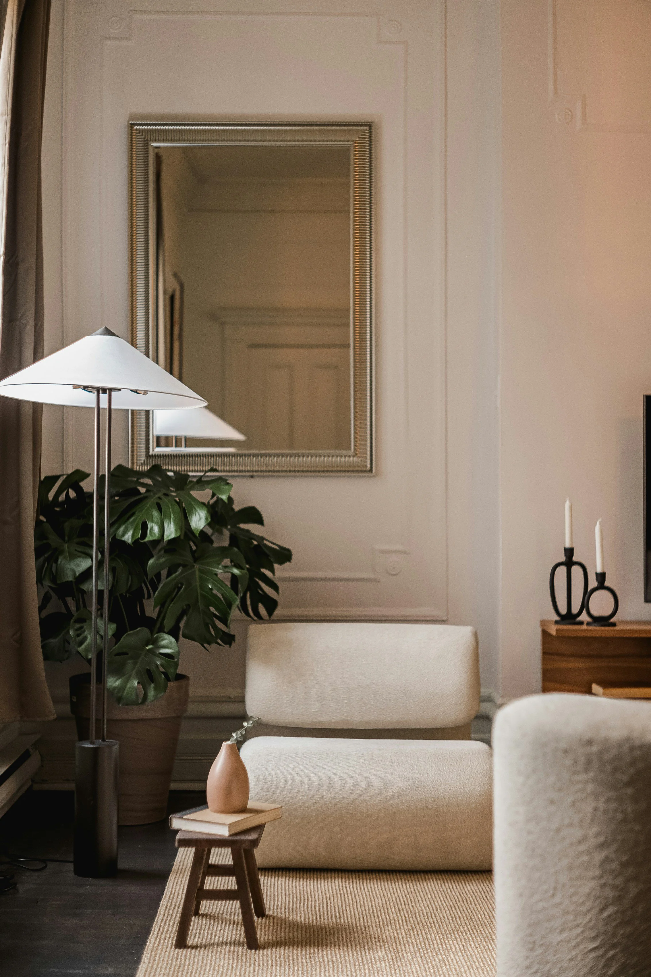 A cozy living room with a beige armchair, a small wooden table with a beige vase and a book, a large potted plant, a floor lamp, a wall mirror, and a wooden side table with two black candle holders and white candles.