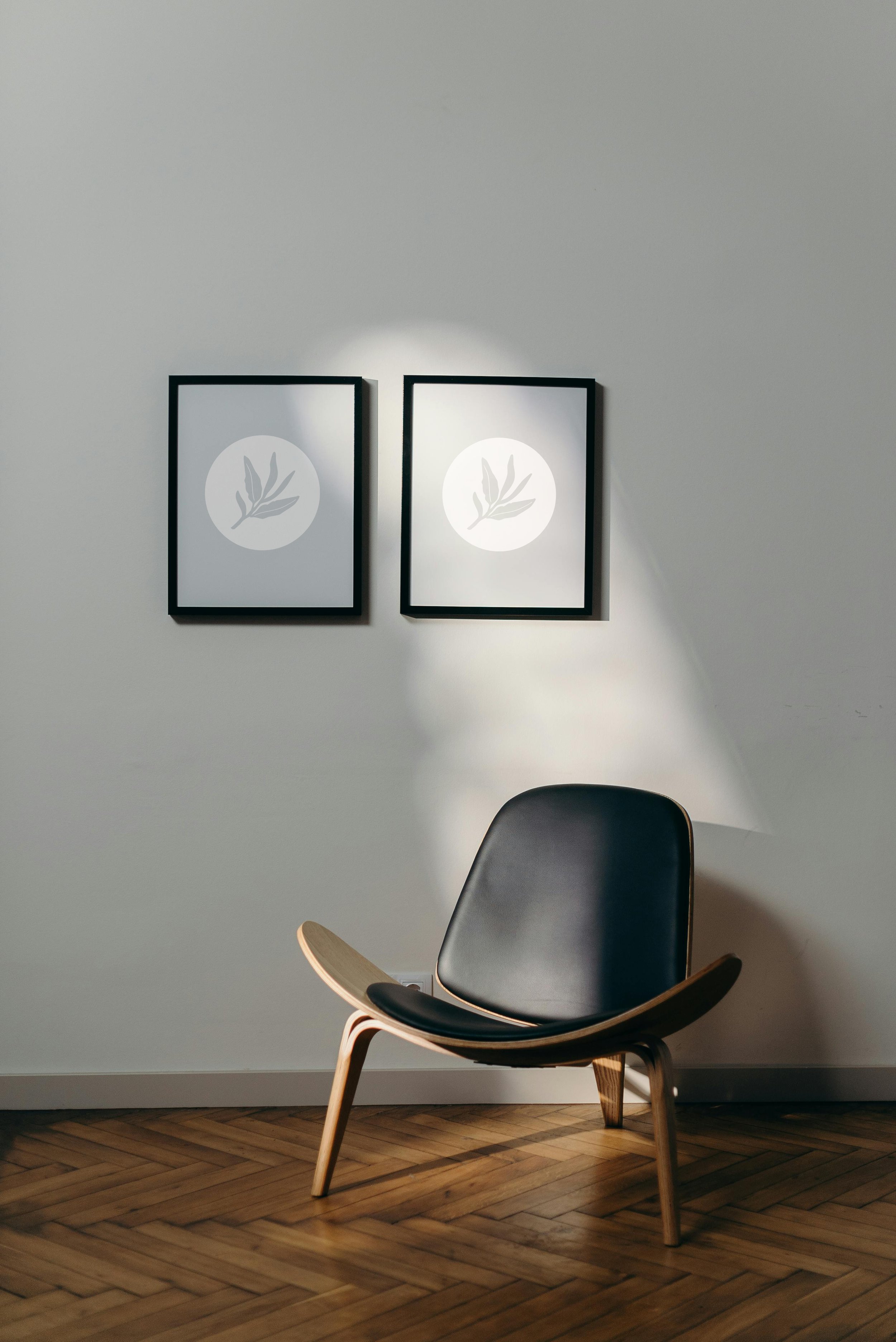 A black and wood lounge chair positioned in a room with hardwood herringbone flooring. Two framed minimalist art pieces with leaf illustrations hang on the white wall behind the chair. Sunlight creates shadows on the wall.