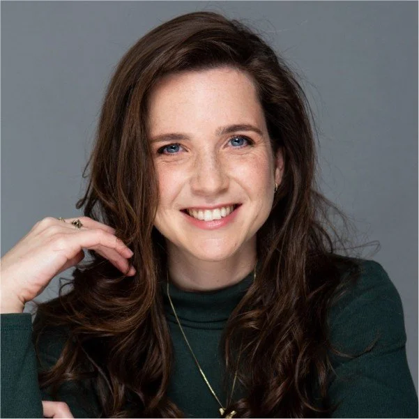 A smiling woman with long brown hair, wearing a dark green top and a gold necklace, poses against a gray background.