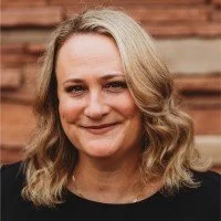 A woman with shoulder-length blonde hair smiling, wearing a black top, standing outdoors with a brick wall in the background.