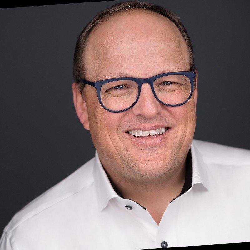 Close-up of a smiling man wearing black-rimmed glasses and a white collared shirt against a dark background.