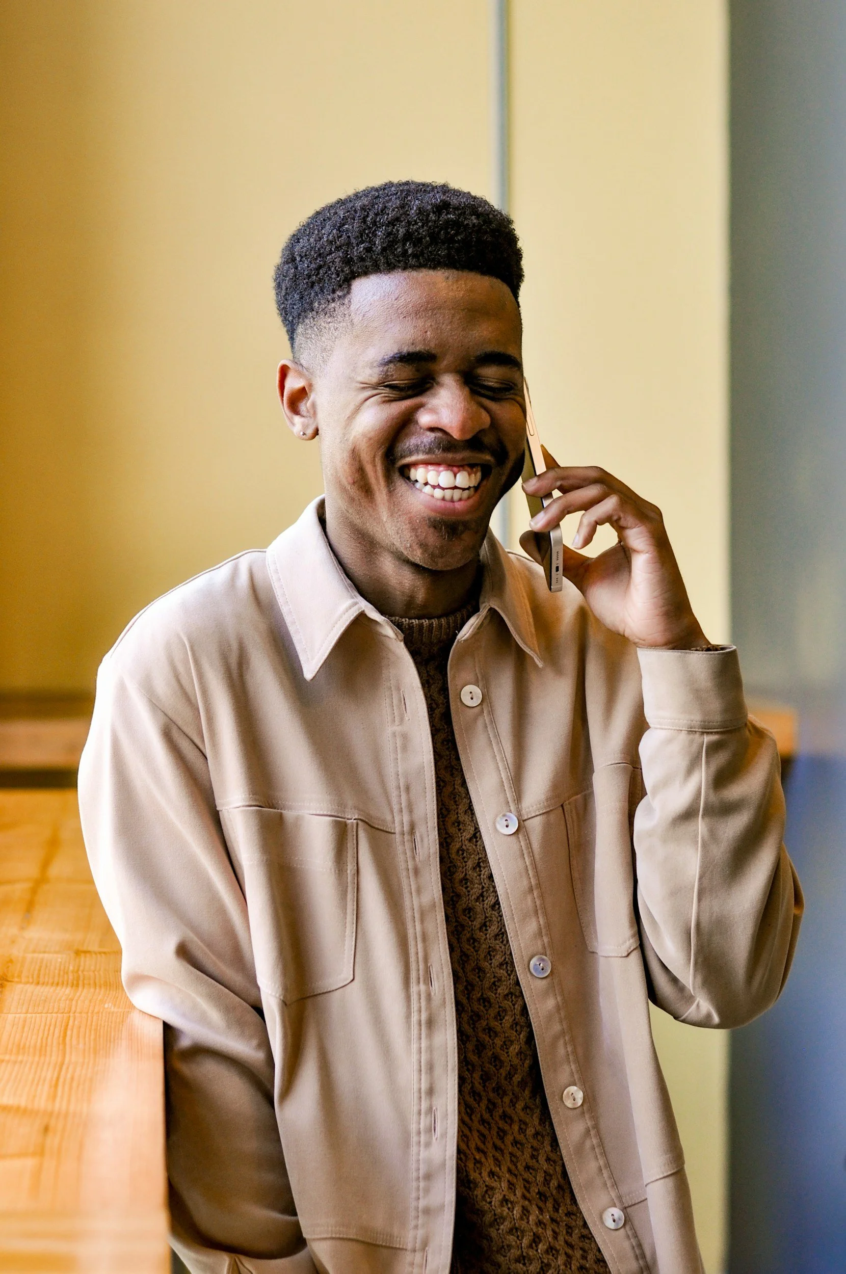 A young man with a short hairstyle smiling and talking on the phone indoors.