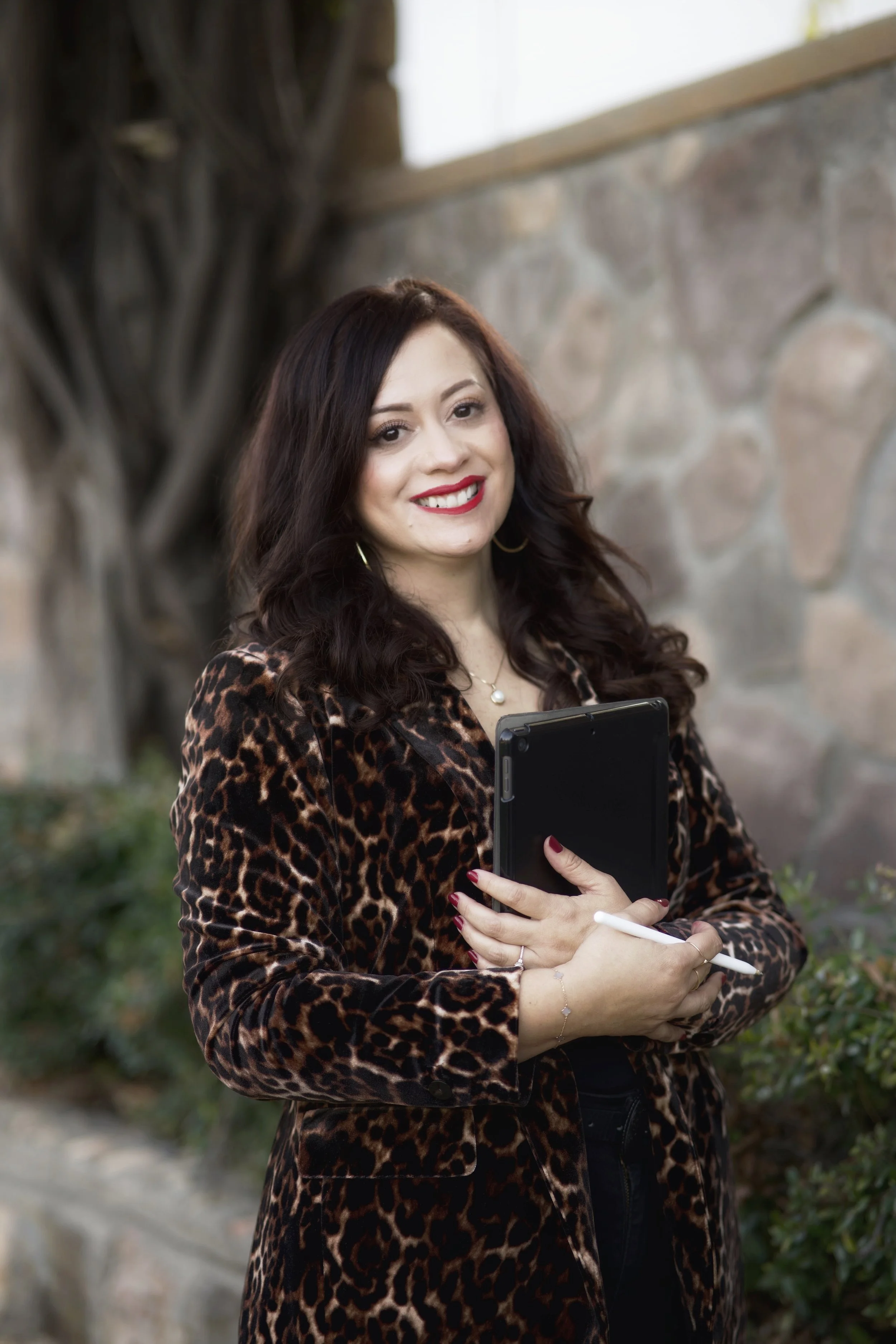 A woman with dark wavy hair, wearing a leopard print jacket and red lipstick, holding a tablet and a stylus, standing outdoors near a stone wall and greenery.