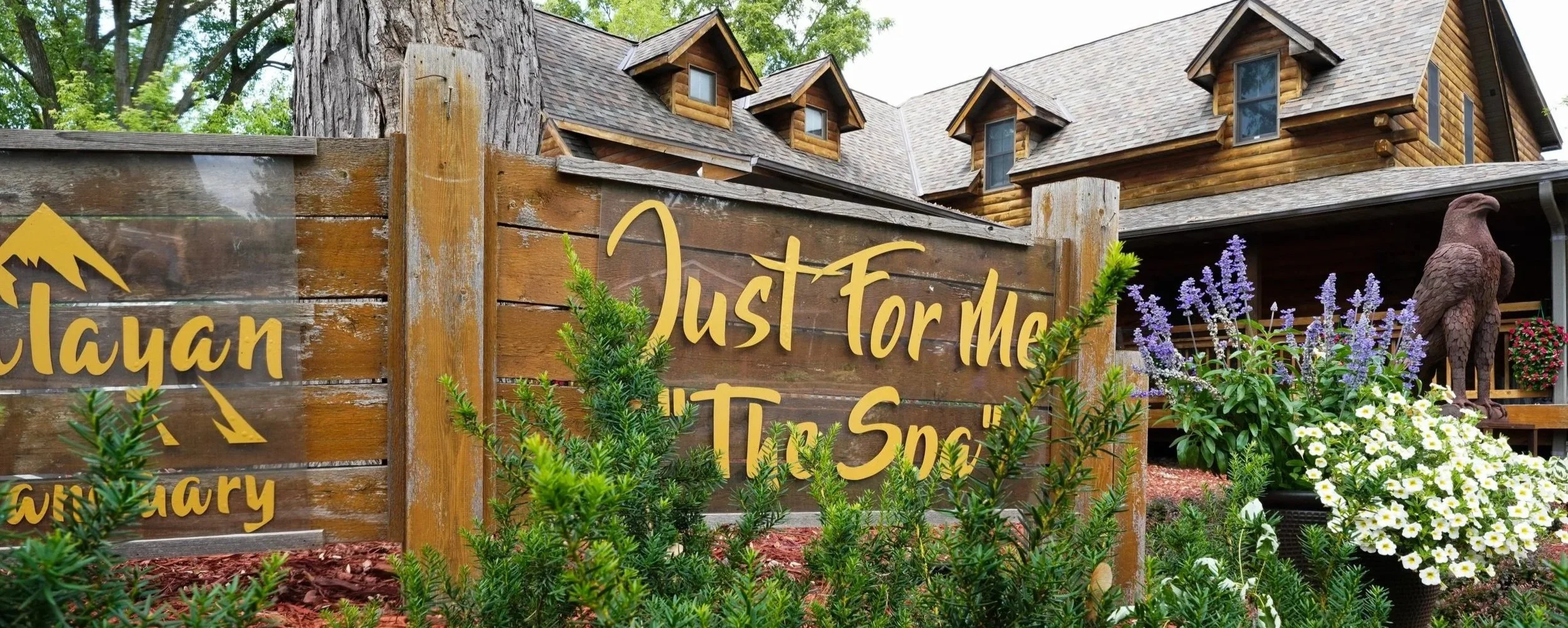 A wooden sign that says 'Just For Me Tlc Spa' next to a wooden fence, with a house, trees, flowers, and a bird-shaped sculpture in the background.