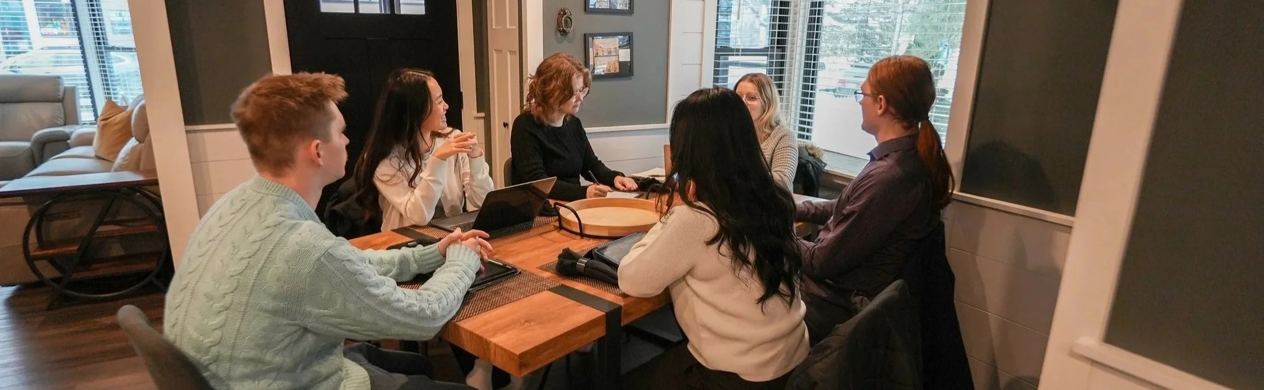 Group of six people sitting around a wooden table in the cozy, well-lit dining room of our Donovan Home, engaged in a conversation or meeting.