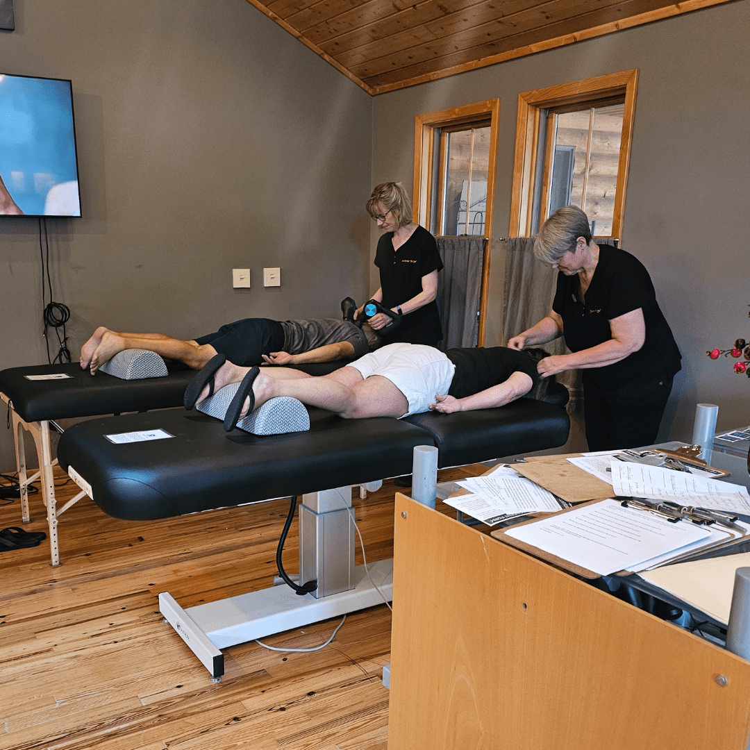 two individuals lying face down on massage table receiving Theragun massage