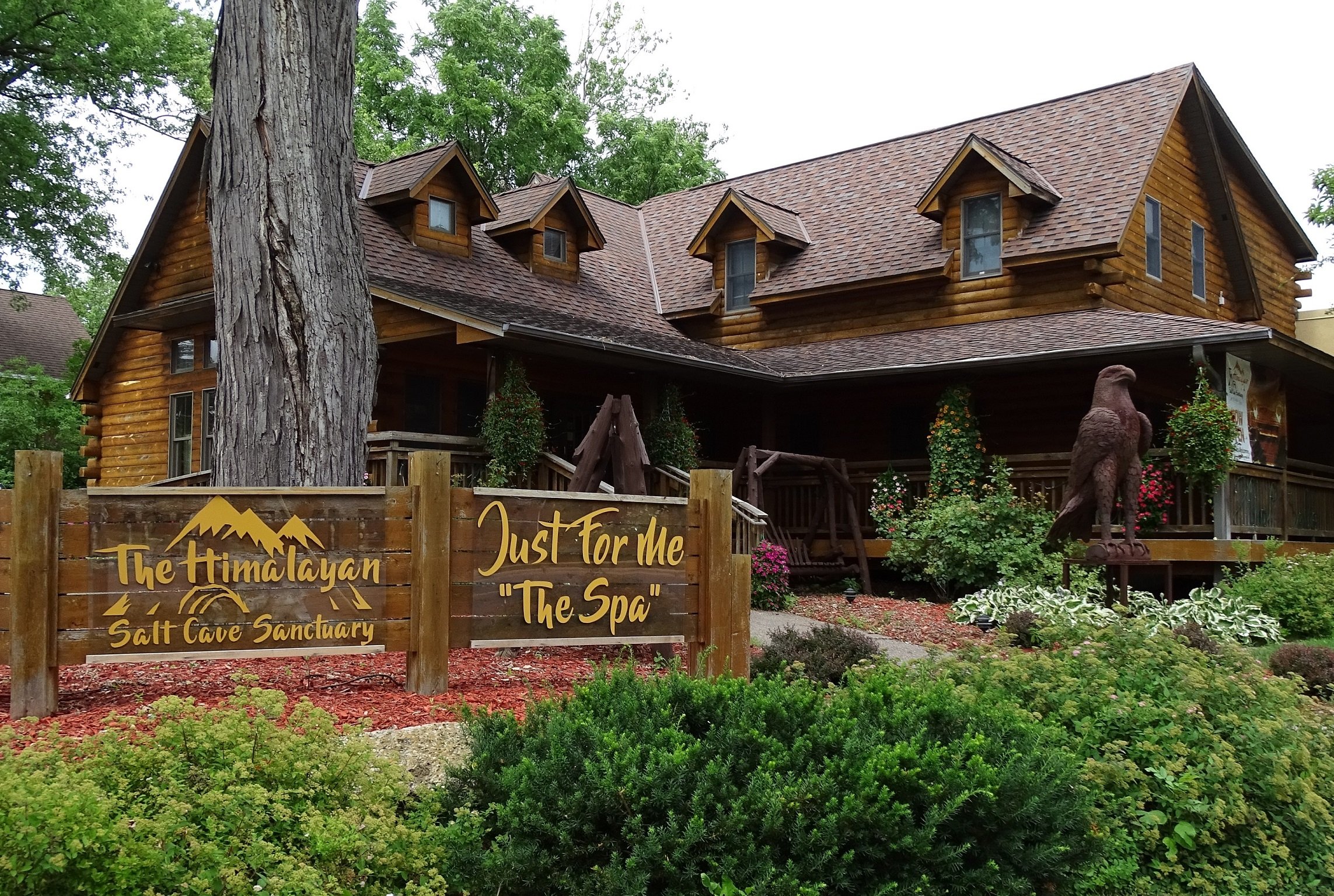 A large wooden building with a steep roof and multiple small dormer windows, surrounded by greenery and flowers, with a wooden sign in front reading 'The Himalayan Salt Cave Sanctuary' and 'Just For Me The Spa'. There is a large eagle statue and a wooden swing near the entrance.