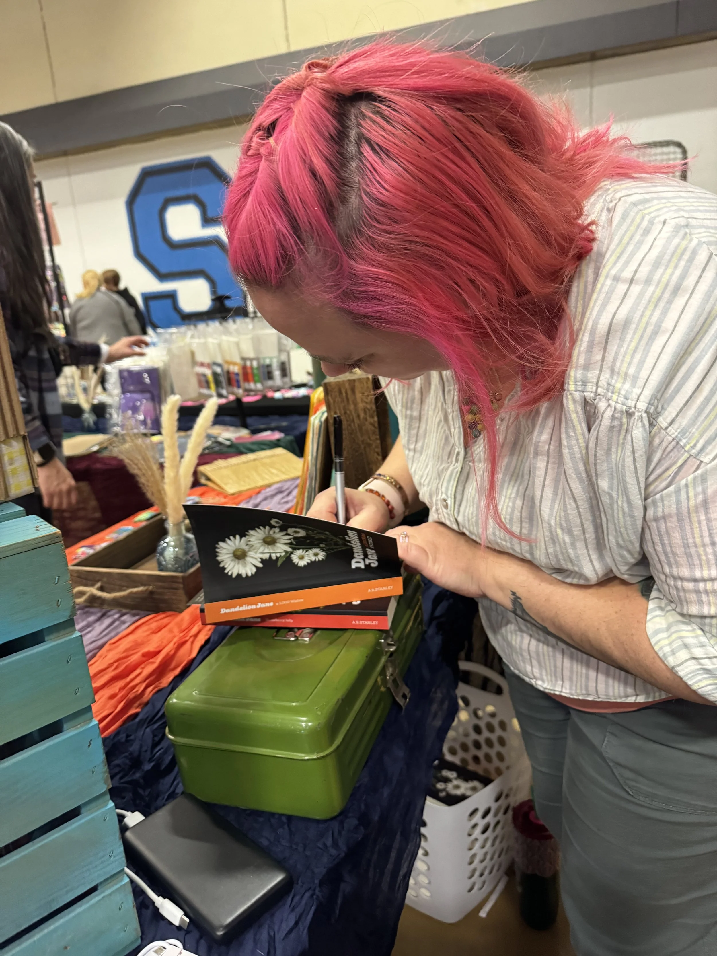 A woman with pink hair signing a book at a table in a crowded indoor event, with various items and a green box on the table.