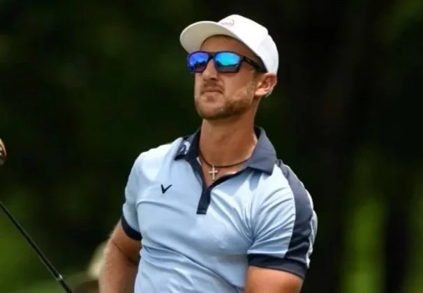 A man is standing on a golf course wearing a white cap, sunglasses, a blue-and-white polo shirt, and a cross necklace. The outdoor golf setting highlights a relaxed, athletic lifestyle.