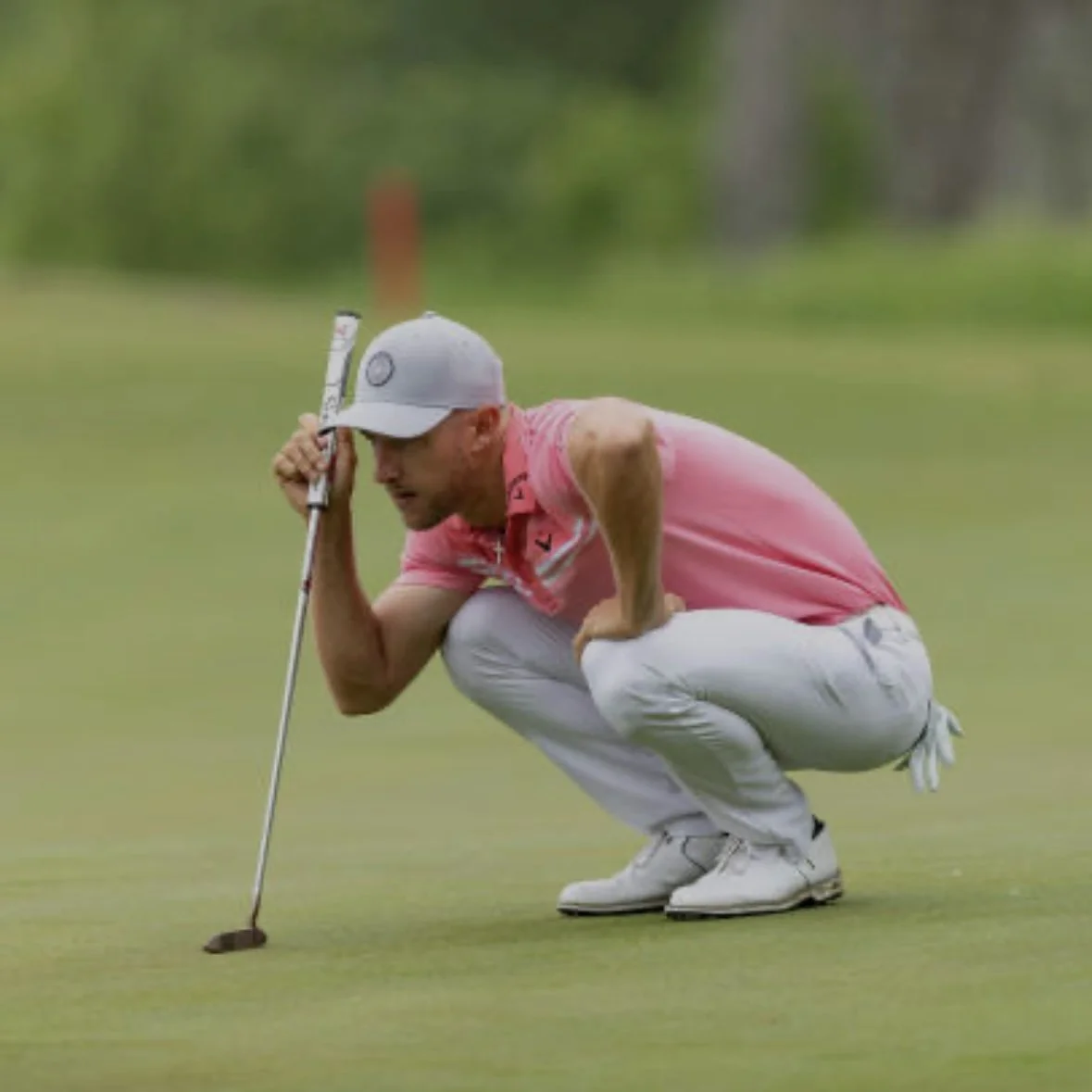 A golfer on the putting green, wearing a pink shirt and gray cap, crouches with a putter, focusing on the shot. This scene highlights putting technique, precision, and sportsmanship, ideal for golf apparel, tips, and lifestyle content.