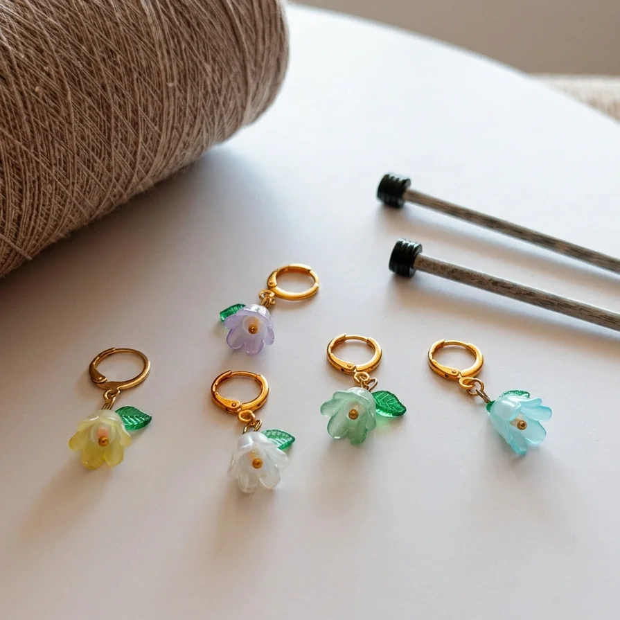 Colorful flower-shaped earrings with green leaves, placed on a white surface near a spool of brown thread and two sewing needles.