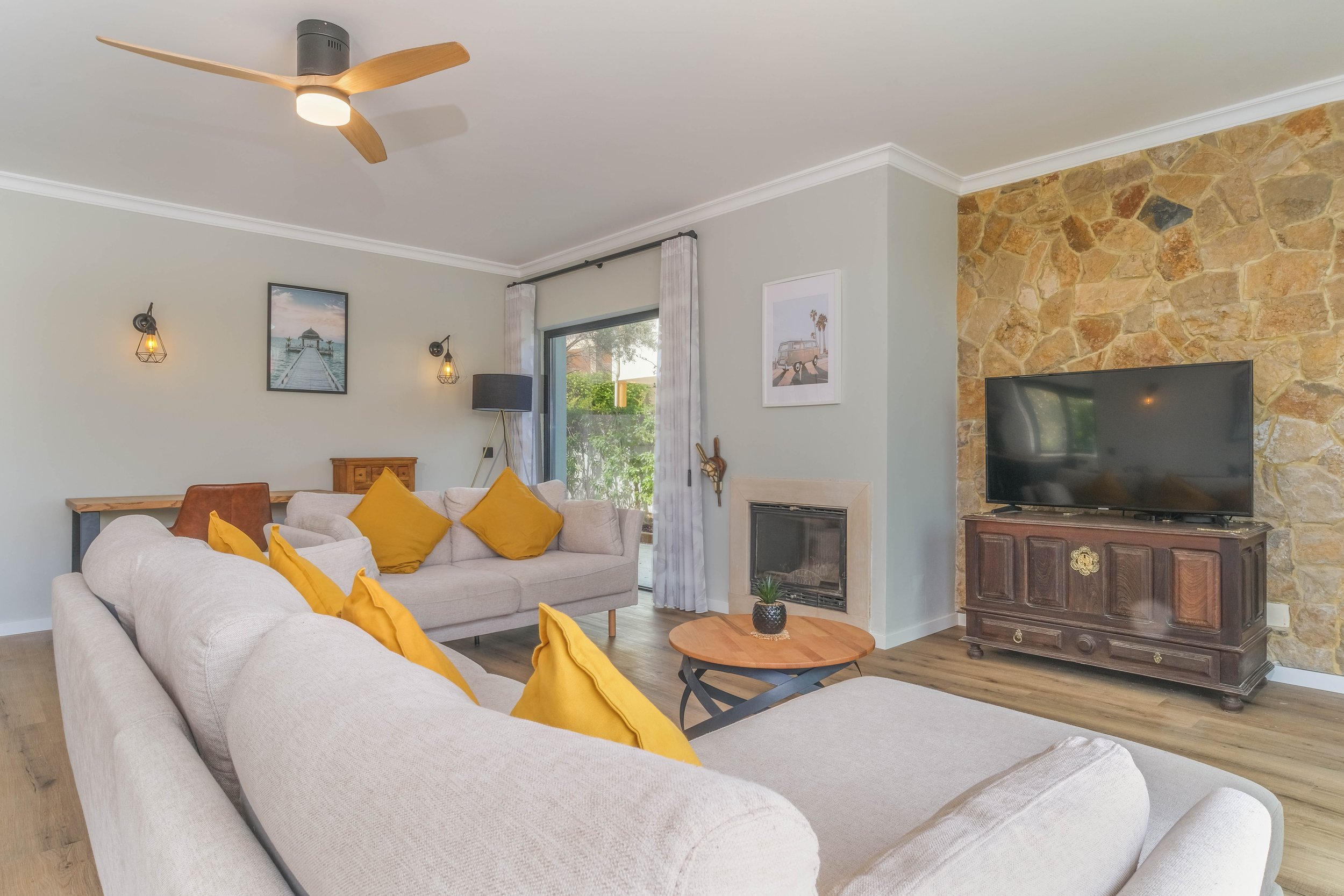 Living room with beige sofas, yellow throw pillows, a wooden coffee table with a plant, stone accent wall with TV, and a sliding glass door with curtains.