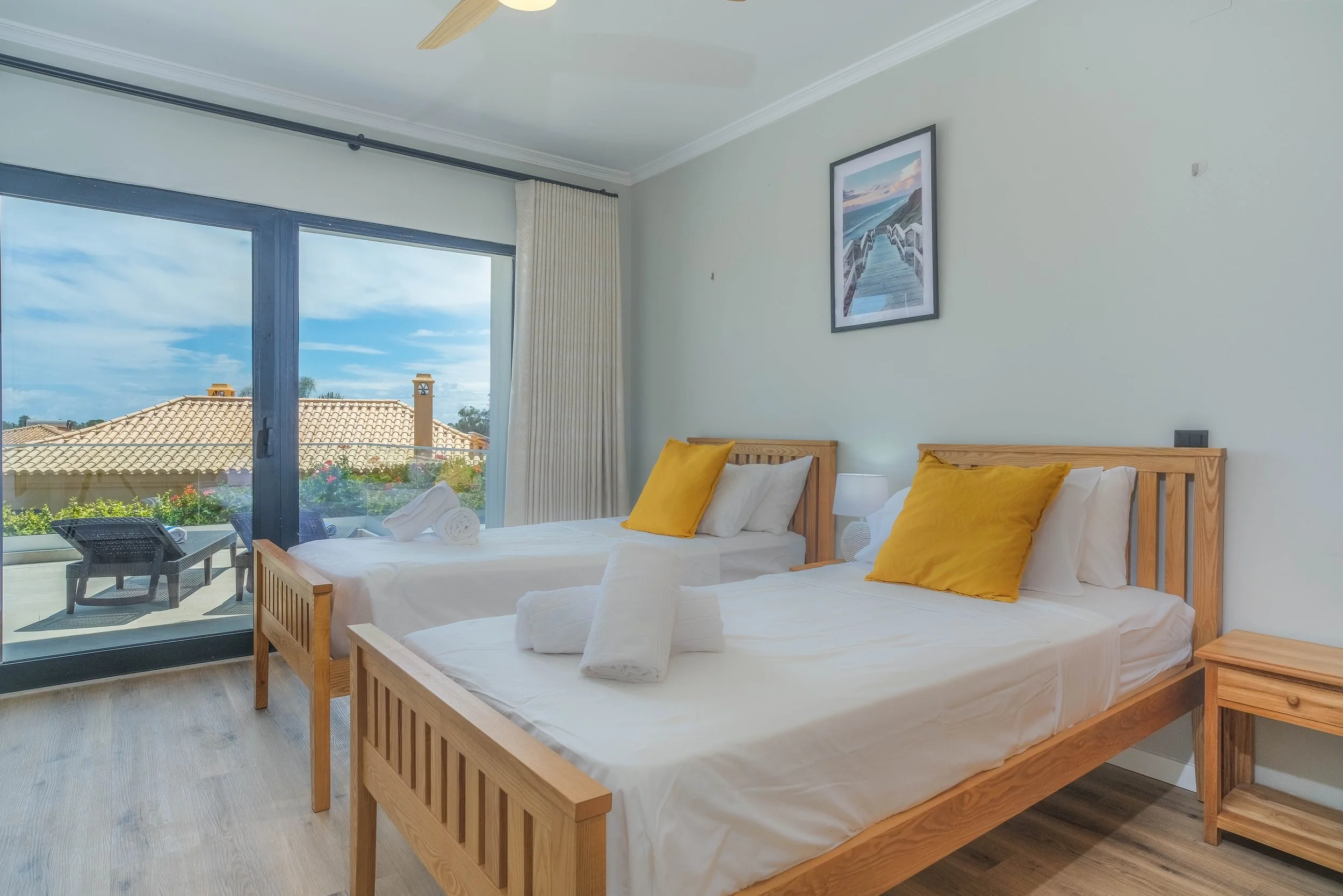 A bedroom with two single beds made of light wood, each with white bedding, white towels, and a mustard yellow pillow. A small wooden nightstand with a lamp is between the beds. There is a large sliding glass door leading to a balcony with outdoor se